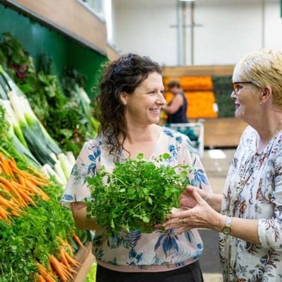 Image of a women with brown curly hair and a lady with short blonde hair smiling and holding green vegetables