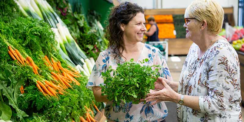 Image of a women with brown curly hair and a lady with short blonde hair smiling and holding green vegetables